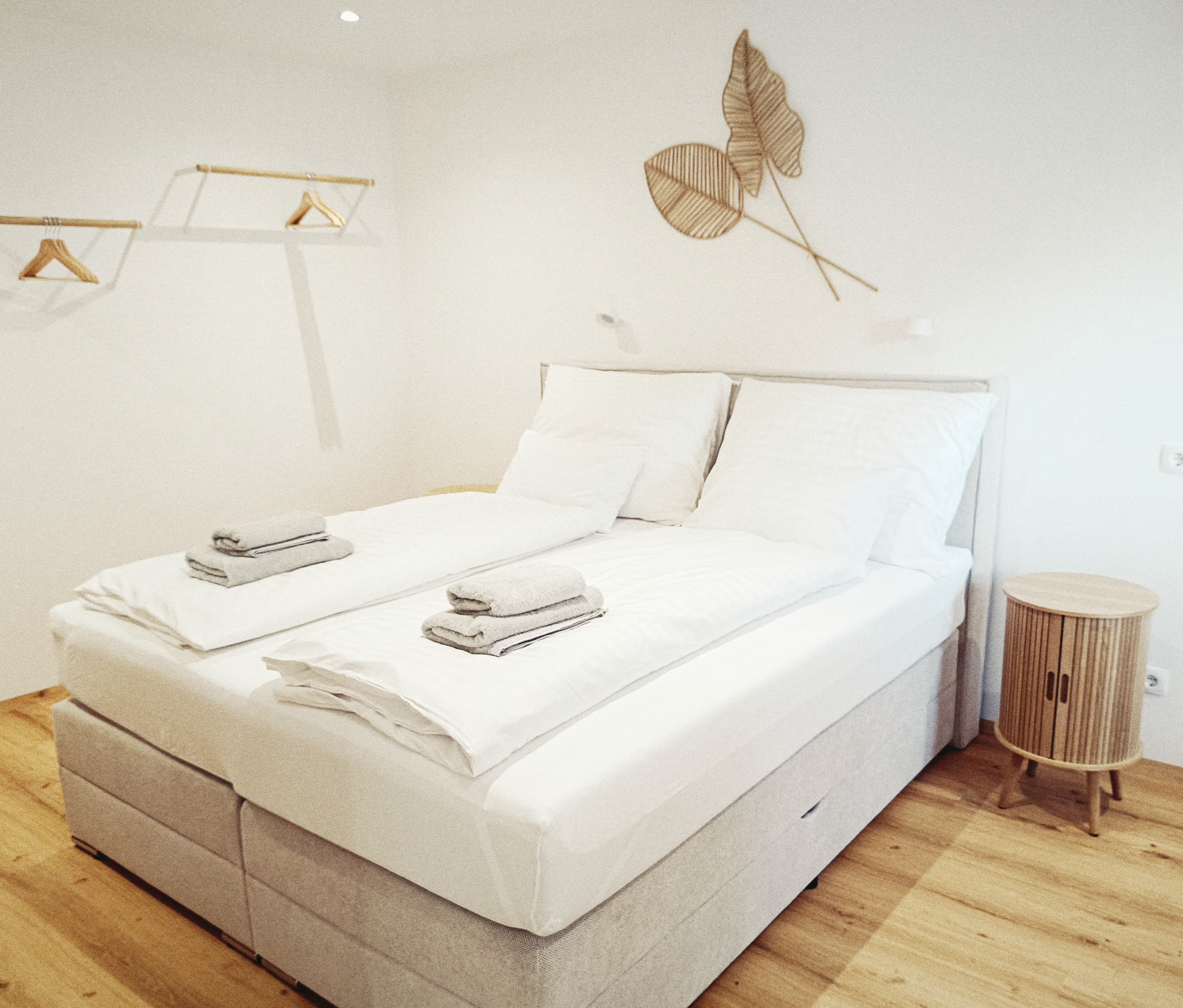 Helles Schlafzimmer mit Doppelbett, weißen Kissen und Handtüchern. Minimalistische Dekoration und Holzboden schaffen eine entspannende Atmosphäre für erholsamen Schlaf.