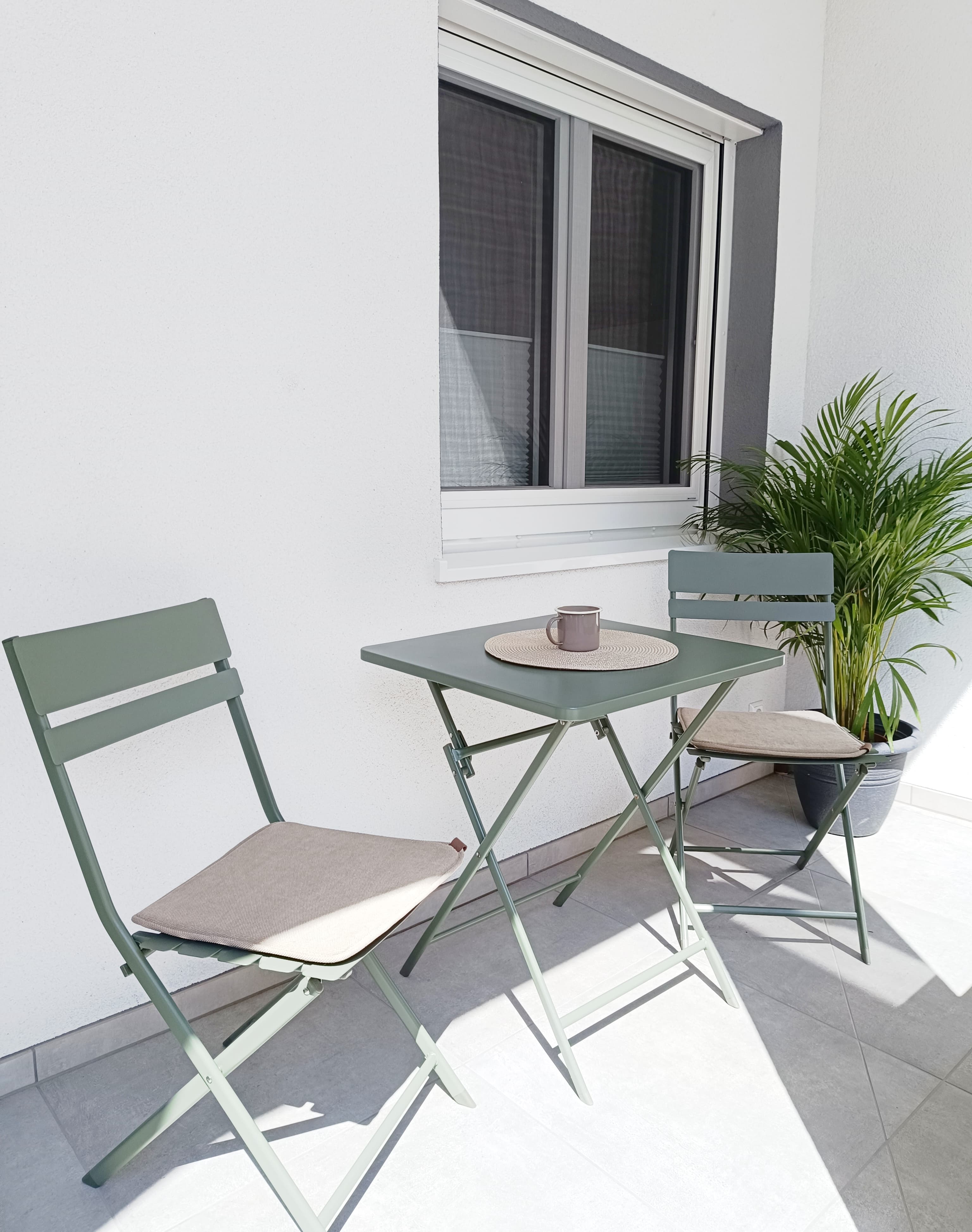 Außenbereich mit Sitzgelegenheiten auf der Terrasse mit einem salbeigrünem Bistrotischset, bestehend aus einem kleinen Tisch, zwei Stühlen mit Kissen und einer Topfpalme. A cozy balcony setup.