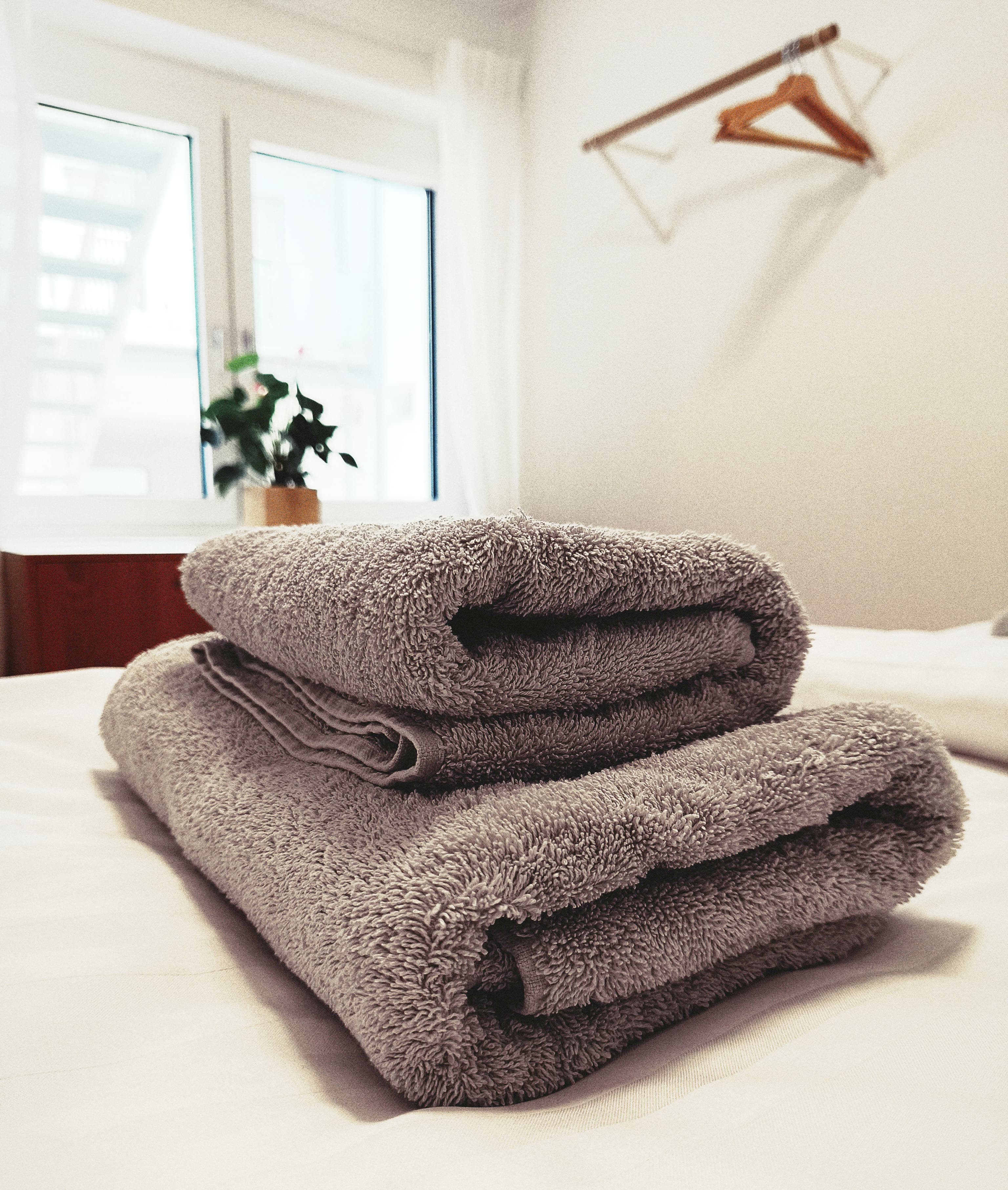 Gemütlicher Stapel gefalteter grauer Handtücher auf einem makellos weißen Bett in einem hellen Raum. Amenities ready for guests. Promoting comfort, relaxation, and hospitality.