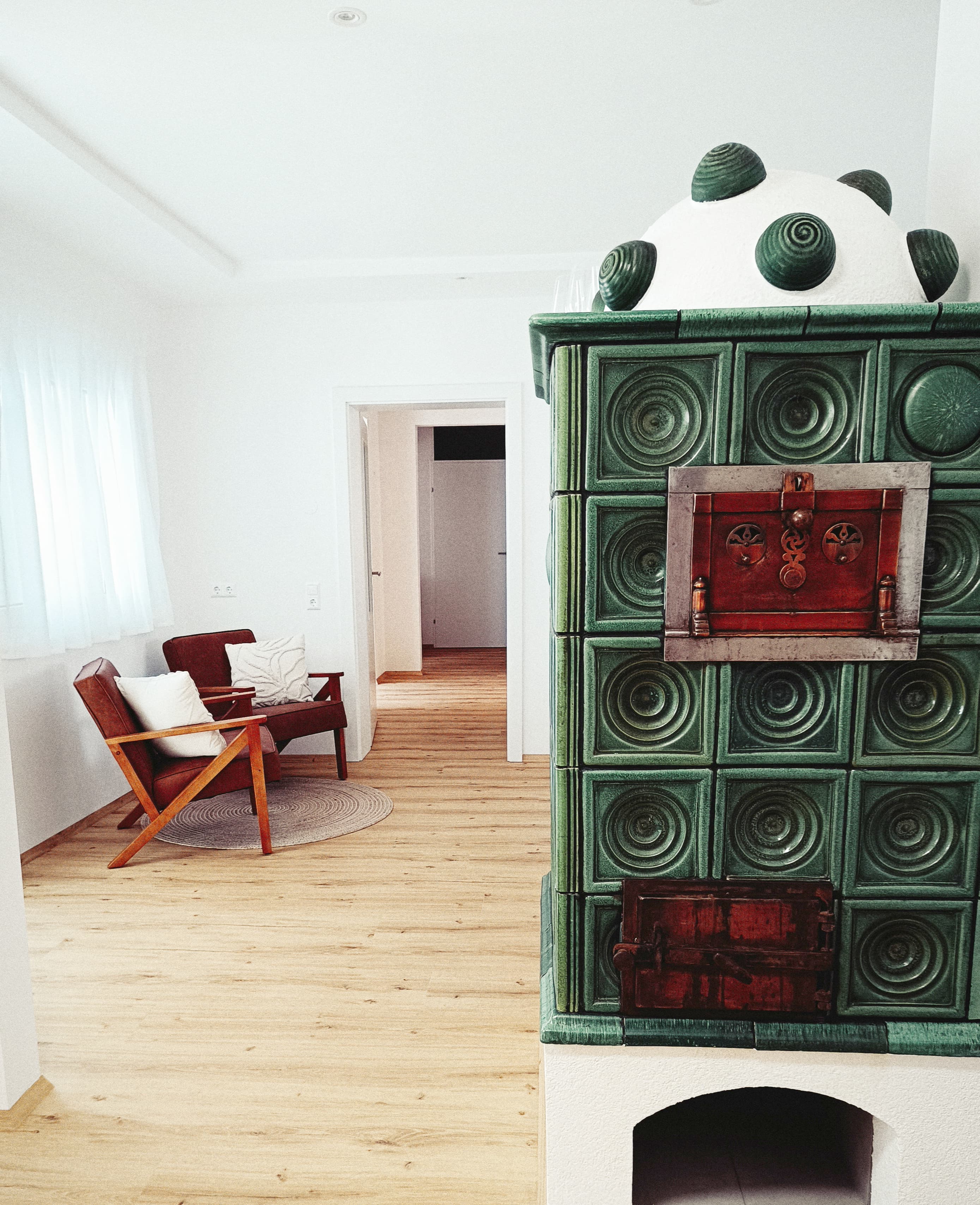 Helles Wohnzimmer mit einem einzigartigen, grünen Kachelofen im Vintage-Stil, zwei modernen braunen Sesseln mit weißen Kissen und hellem Holzboden für eine gemütliche Atmosphäre.