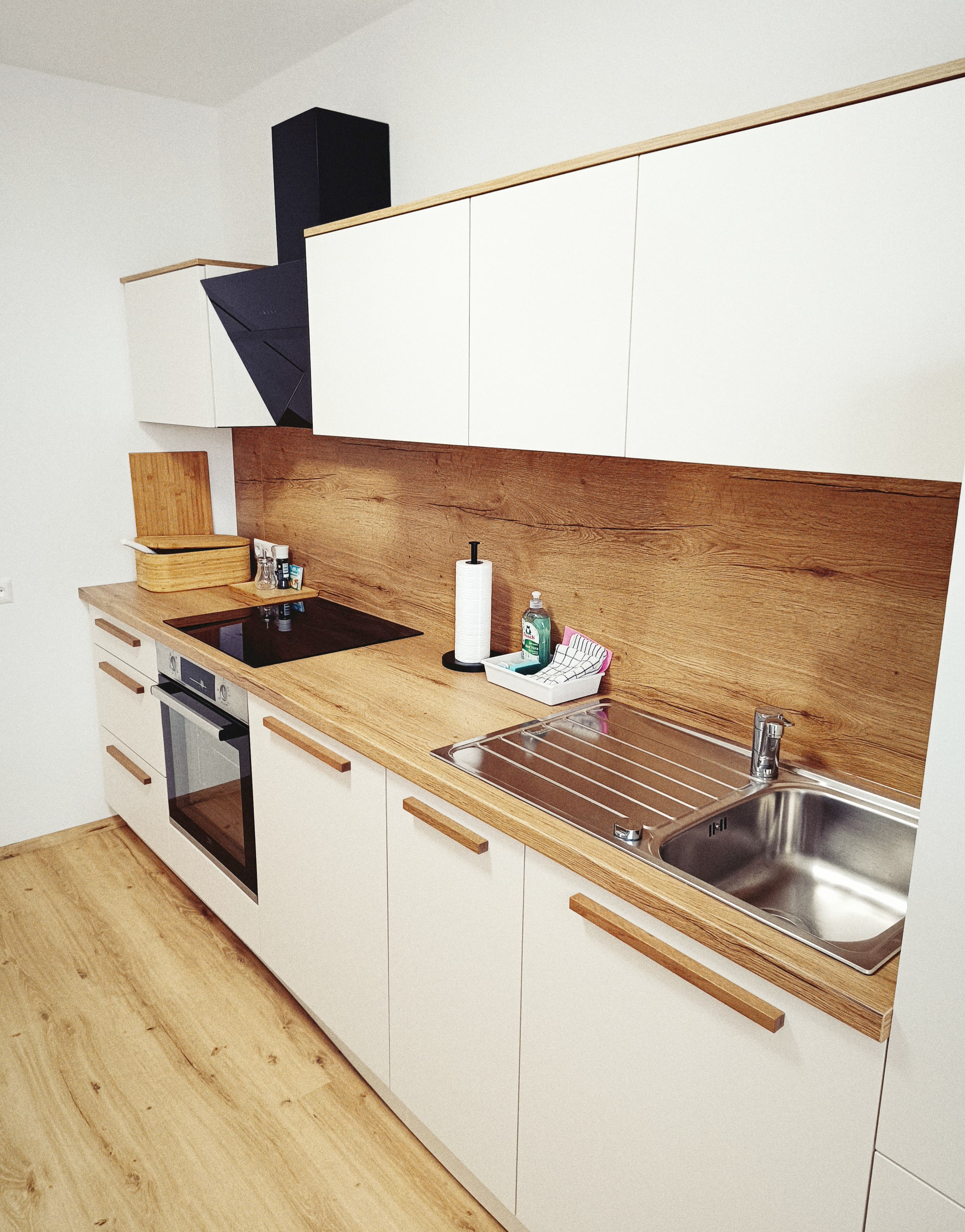 Moderne Kücheneinrichtung mit weißen Schränken, einer Arbeitsplatte mit Holzmaserung, einem Edelstahlspülbecken und minimalistischen Holzgriffen. It's well-lit and stylish.