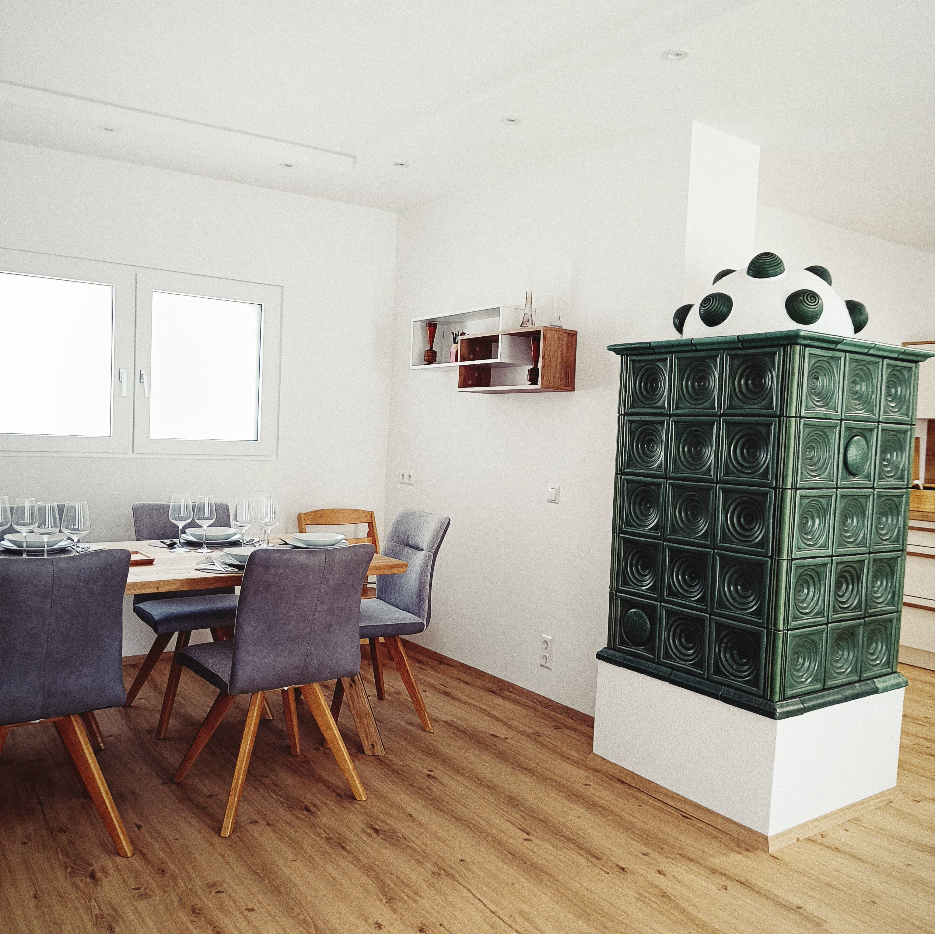 Helles, luftiges Esszimmer mit einem langen Holztisch, der für ein Mahl gedeckt ist, modernen grauen Stühlen und einem einzigartigen grünen Kachelofen in der Ecke.