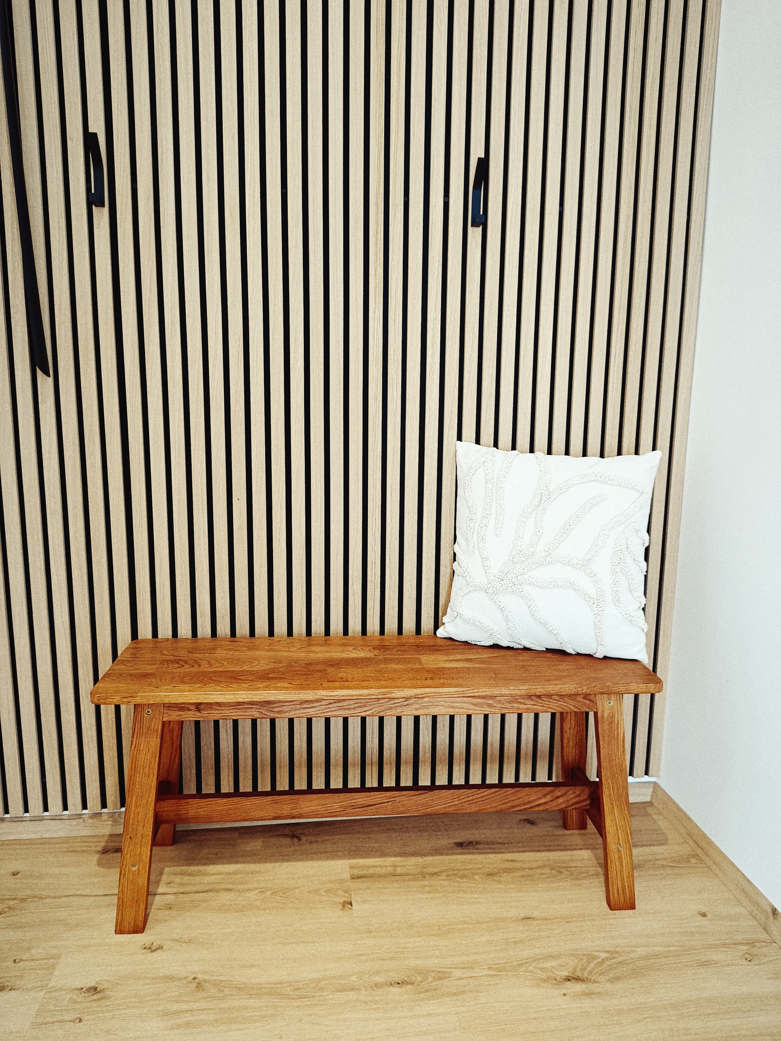 Eine Holzbank mit einem weißen Kissen steht an einer Wand mit vertikalen schwarzen Streifen und schafft eine minimalistische und ruhige Atmosphäre.