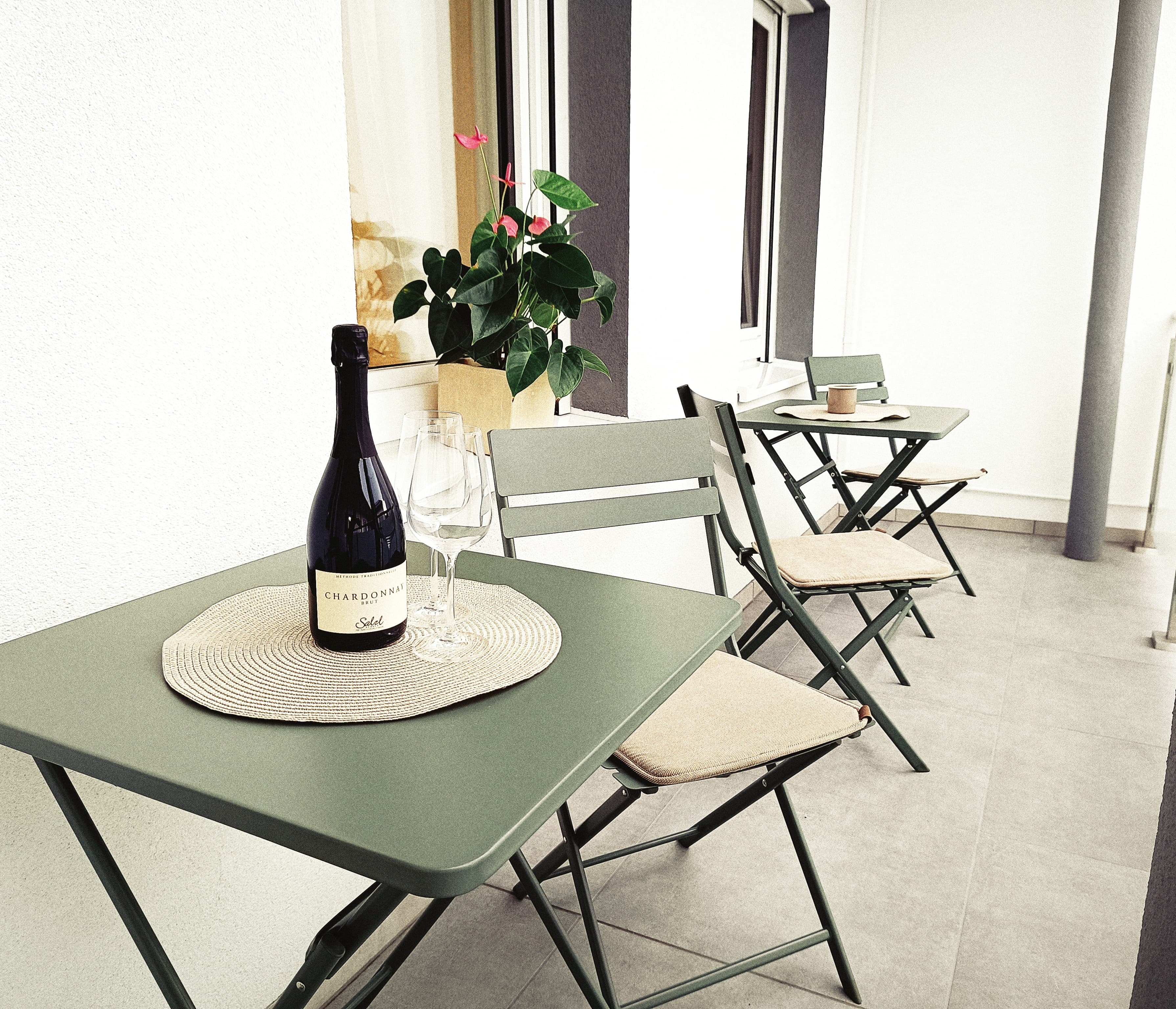 Gemütliche Balkonszene mit grünen Tischen und Stühlen. A bottle of wine and glasses sit on a placemat. A potted plant adds a touch of nature.