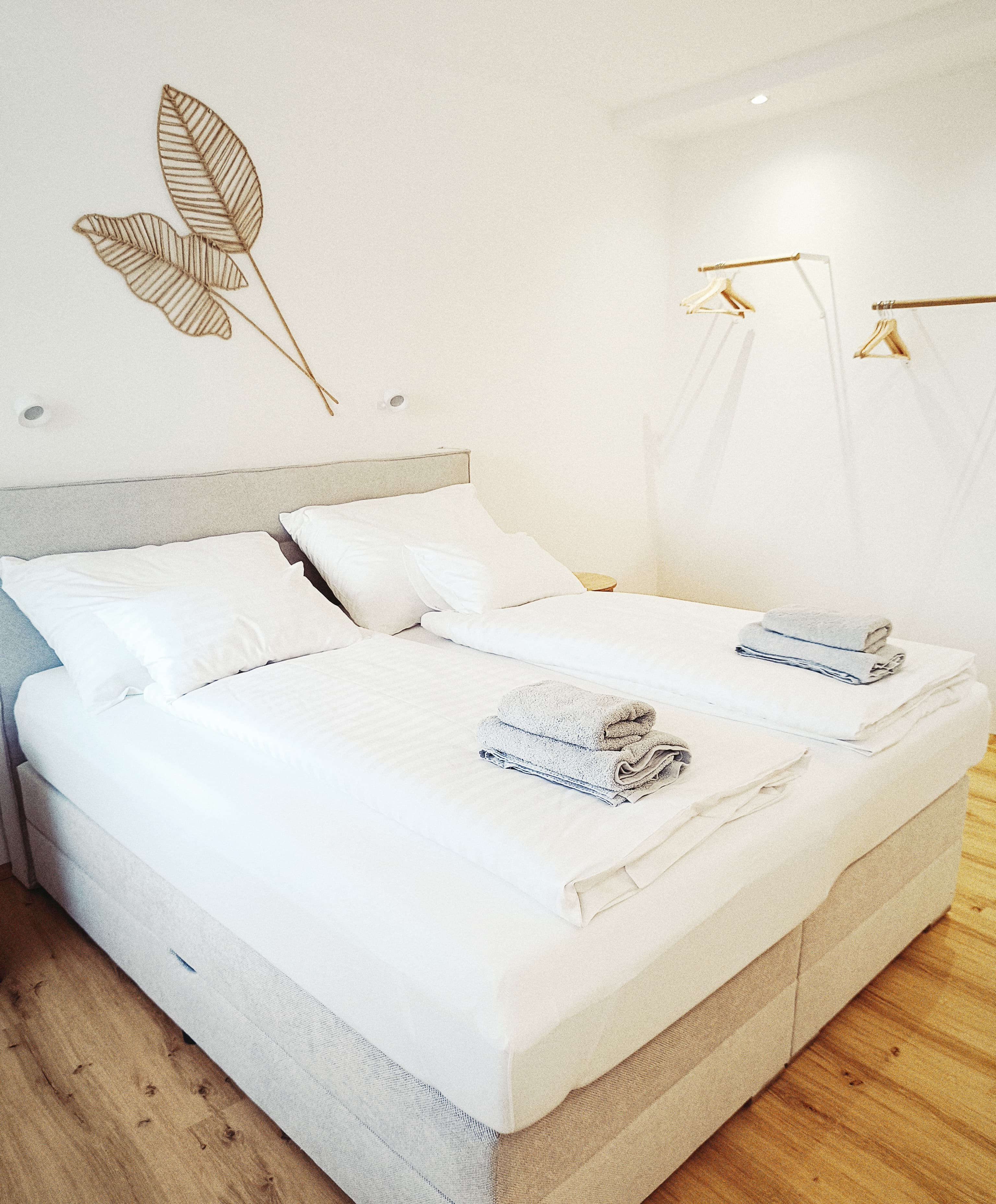 Minimalistisches Schlafzimmer mit zwei ordentlich gemachten Einzelbetten, weißer Bettwäsche, gefalteten grauen Handtüchern, Holzboden, weißen Wänden. Leaf wall art and simple wooden hangers create a calm, airy feel.