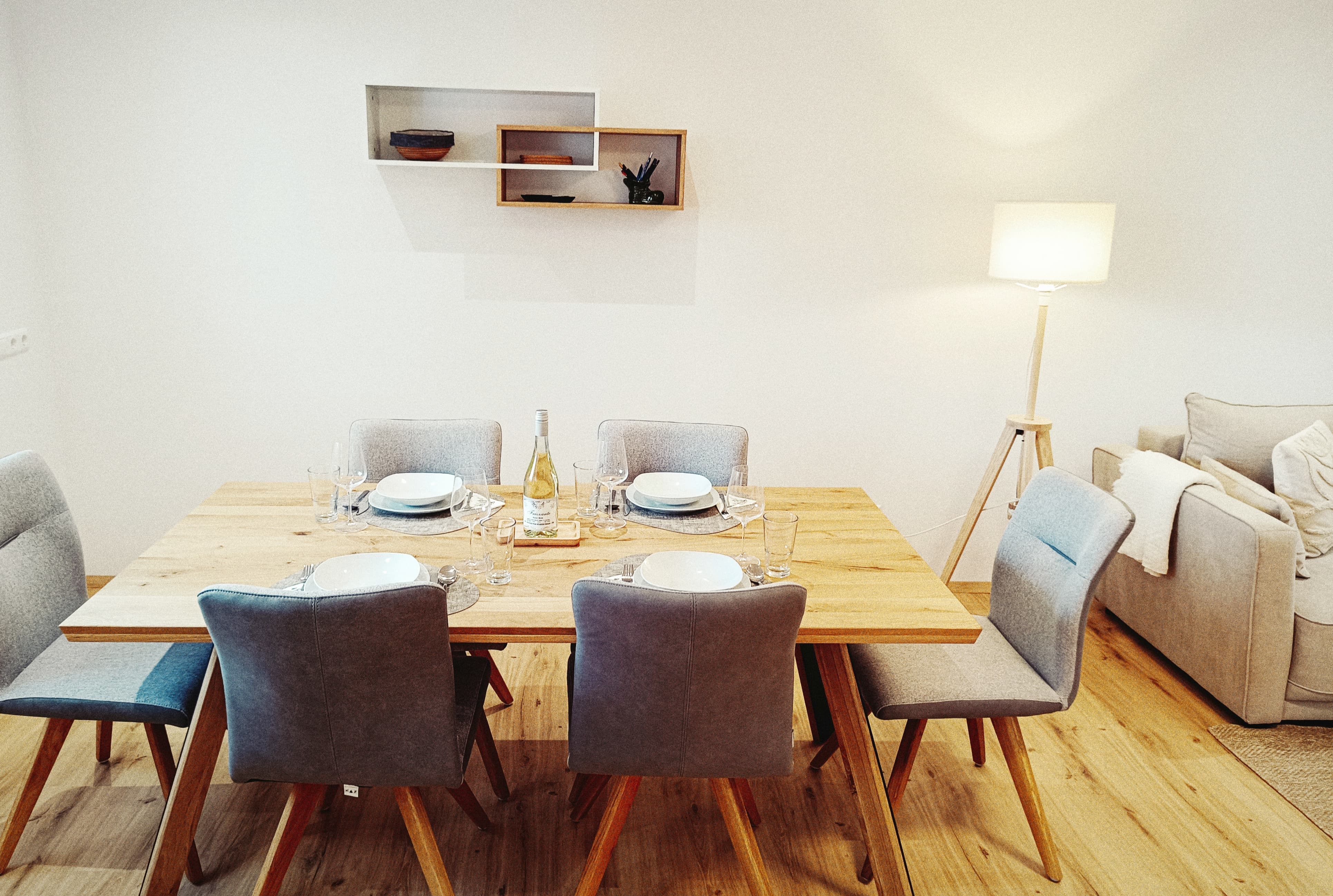 Speisesaal mit einem Holztisch für sechs Personen, ausgestattet mit grauen Stühlen, weißen Tellern und einer Weinflasche. A cozy ambiance with a floor lamp and sofa.
