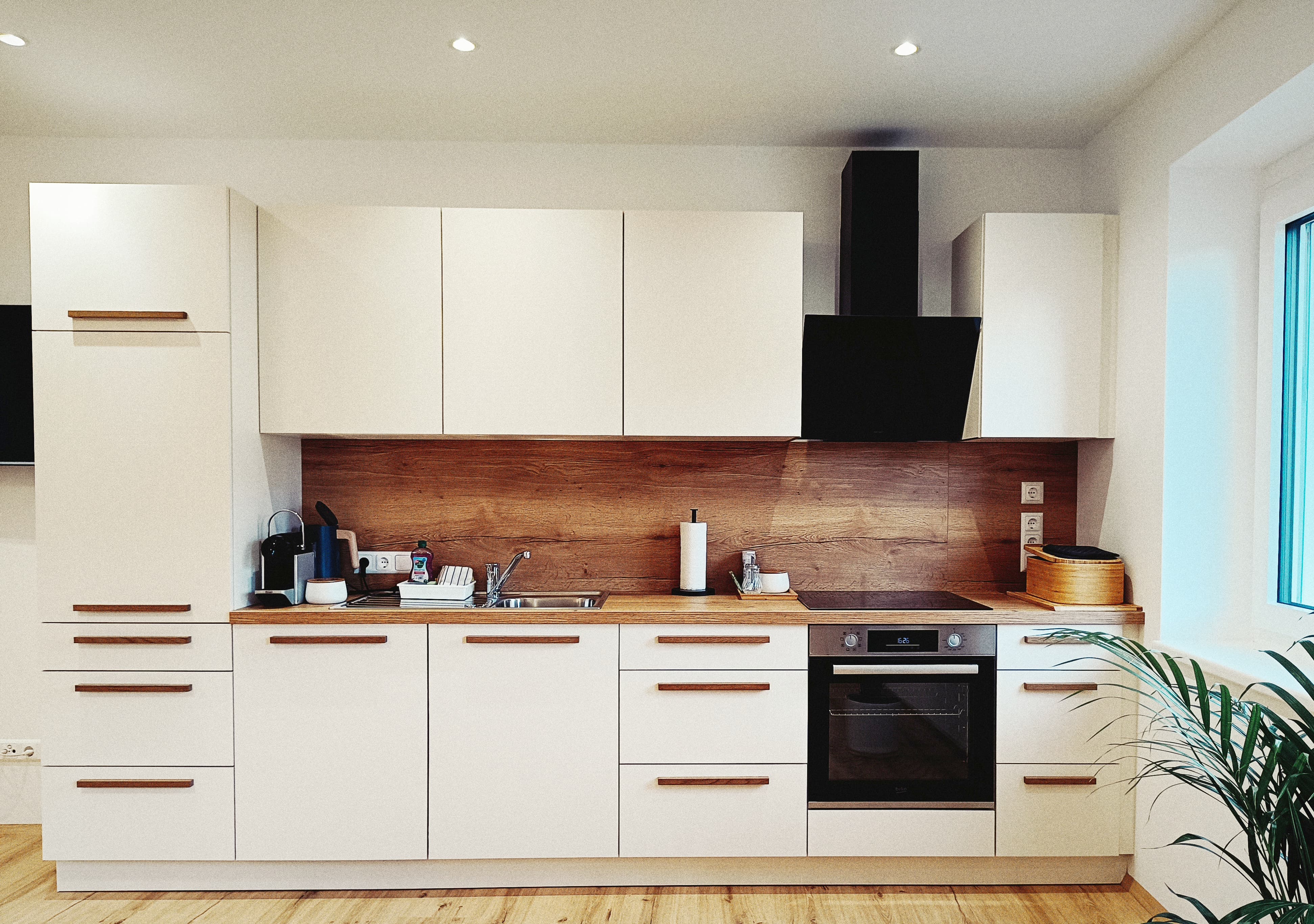 Moderne Küche mit weißen Schränken, Arbeitsplatte aus Holz und einem schwarzen Backofen. Wooden backsplash adds warmth. Nearby window and plant enhance brightness.