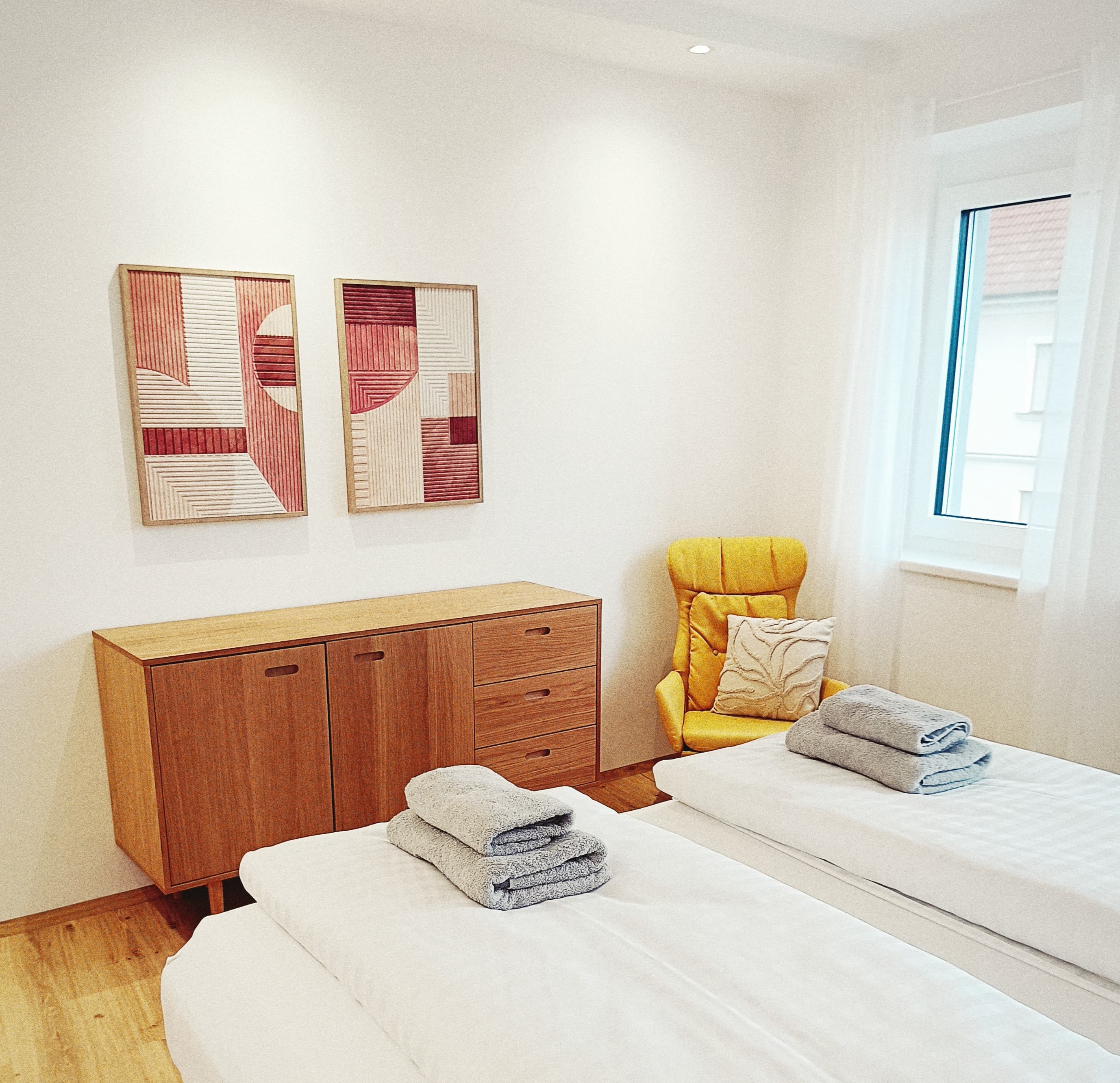 Ein helles, minimalistisches Schlafzimmer mit zwei weißen Betten, auf jedem liegen gefaltete graue Handtücher. A wooden sideboard holds abstract art above it. A yellow chair is by a window.