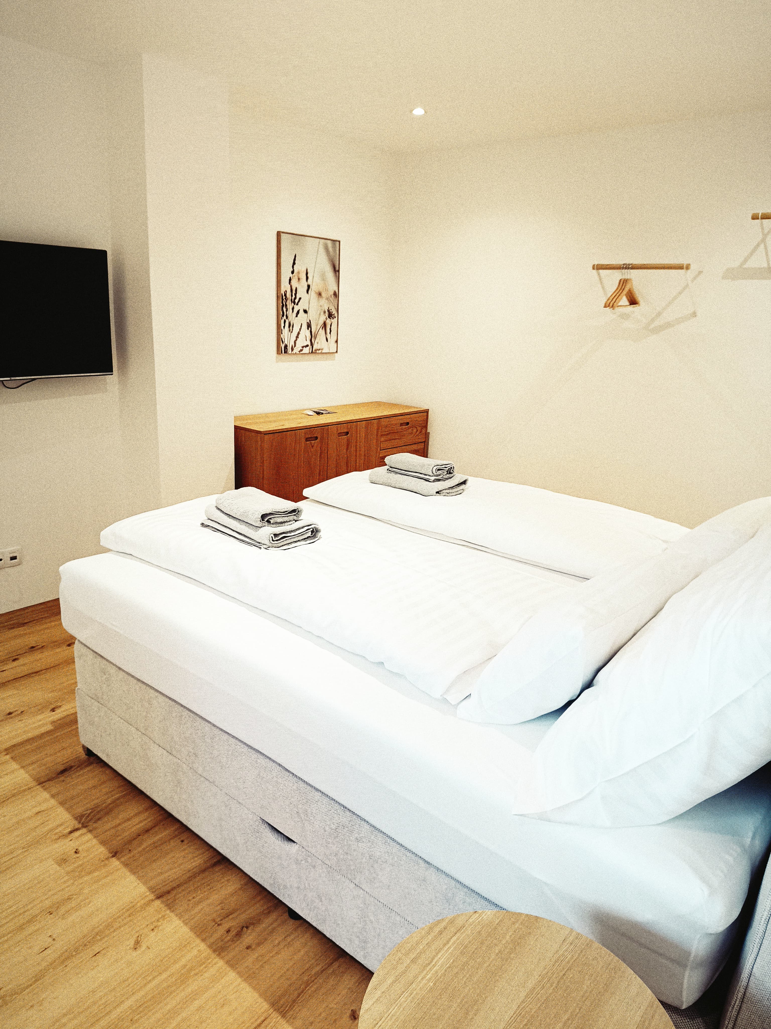 Ein gemütliches, minimalistisches Schlafzimmer verfügt über ein ordentlich gemachtes Doppelbett mit weißer Bettwäsche und gefalteten Handtüchern. A TV is mounted on the wall, above a wooden cabinet.