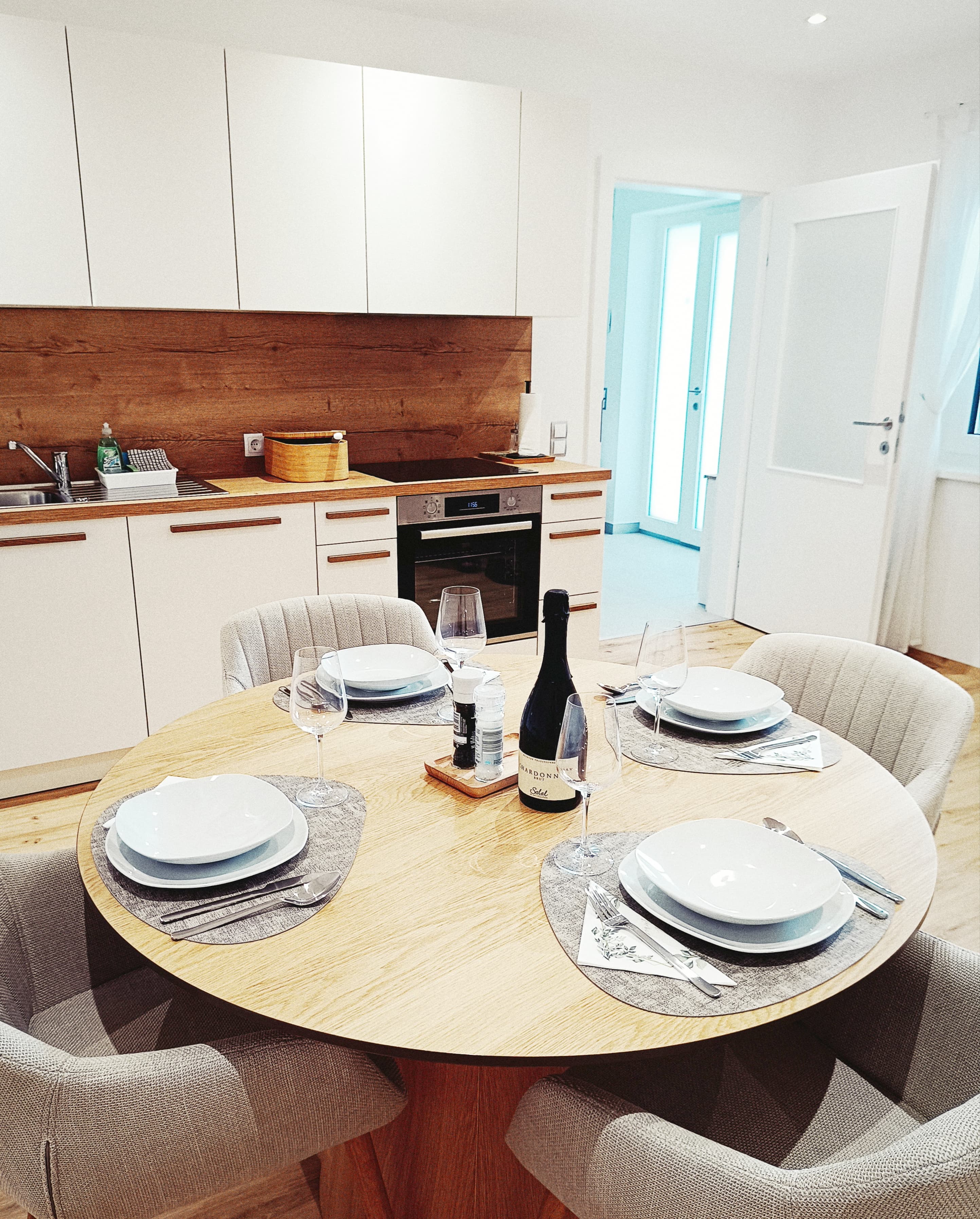 Modern kitchen with a wooden dining table set for four, featuring white plates, wine glasses, and a bottle. Light, inviting ambiance with beige tones.