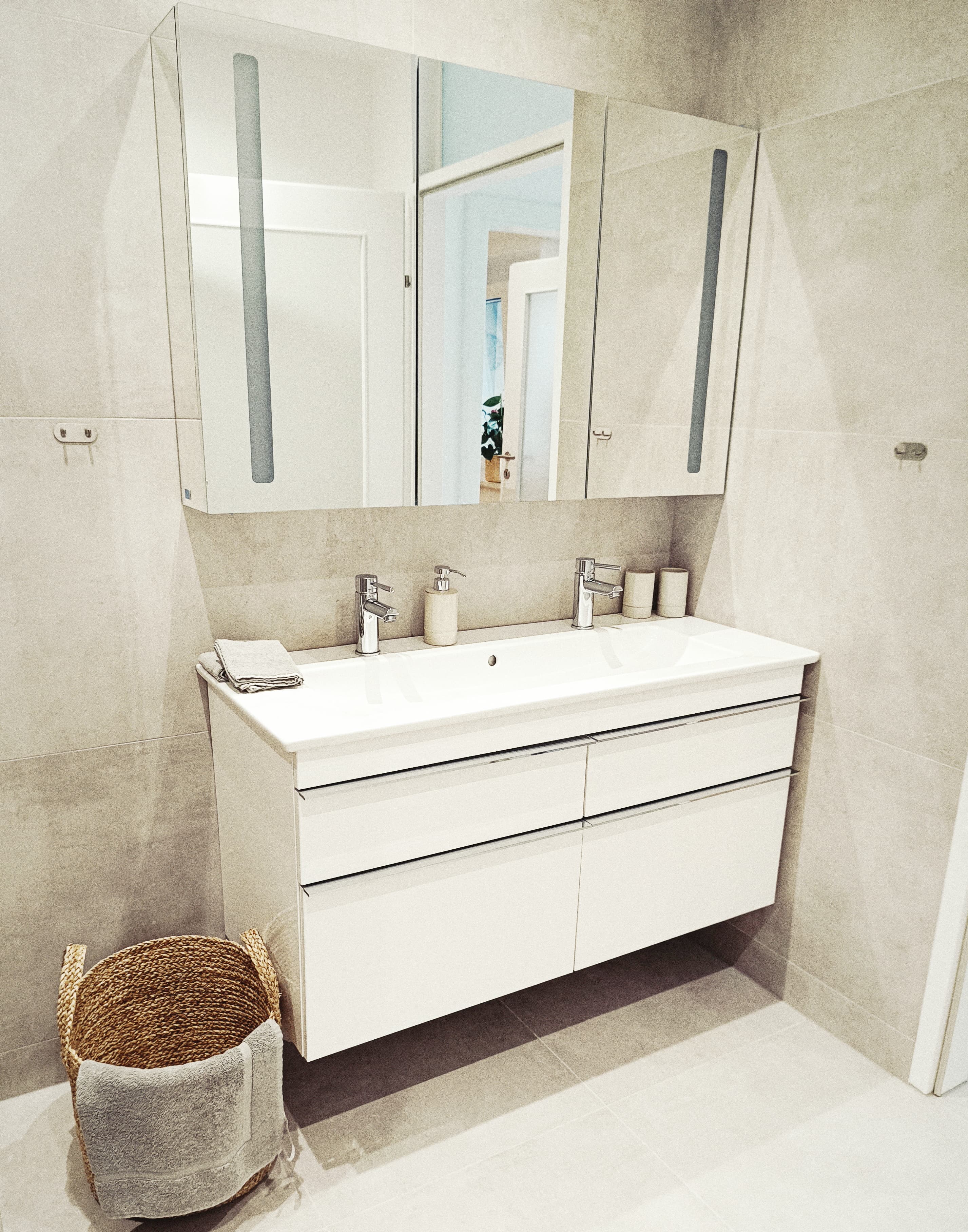 Modernes Badezimmer mit einem eleganten weißen Doppelwaschtisch und großem Spiegel. Neutral tones, clean design, and wicker basket create a calm, minimalist ambiance.