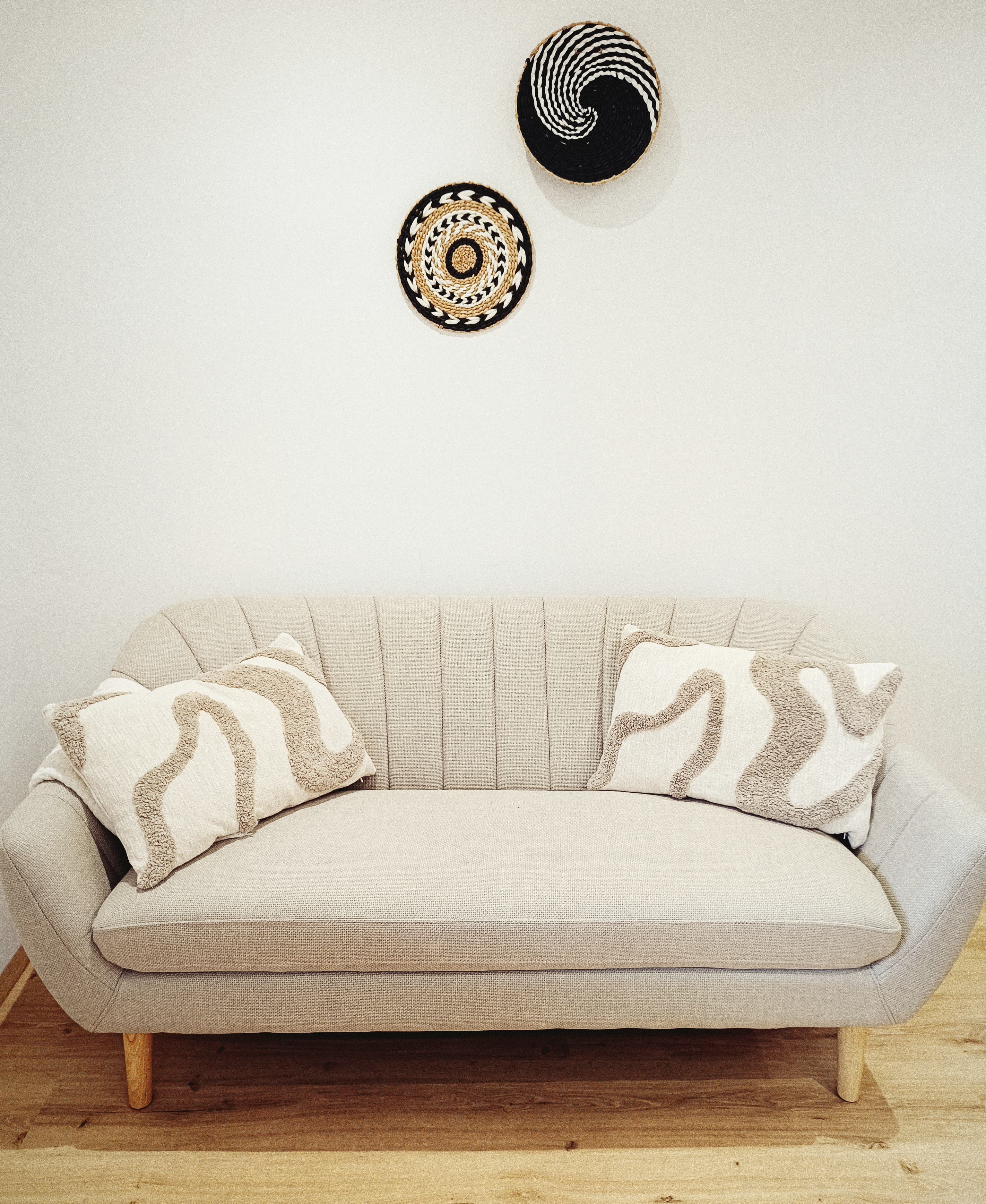 Ein beigefarbenes, texturiertes Sofa mit zwei weißen Kissen mit abstrakten Mustern steht vor einer weißen Wand. Above, two artistic woven baskets hang decoratively.