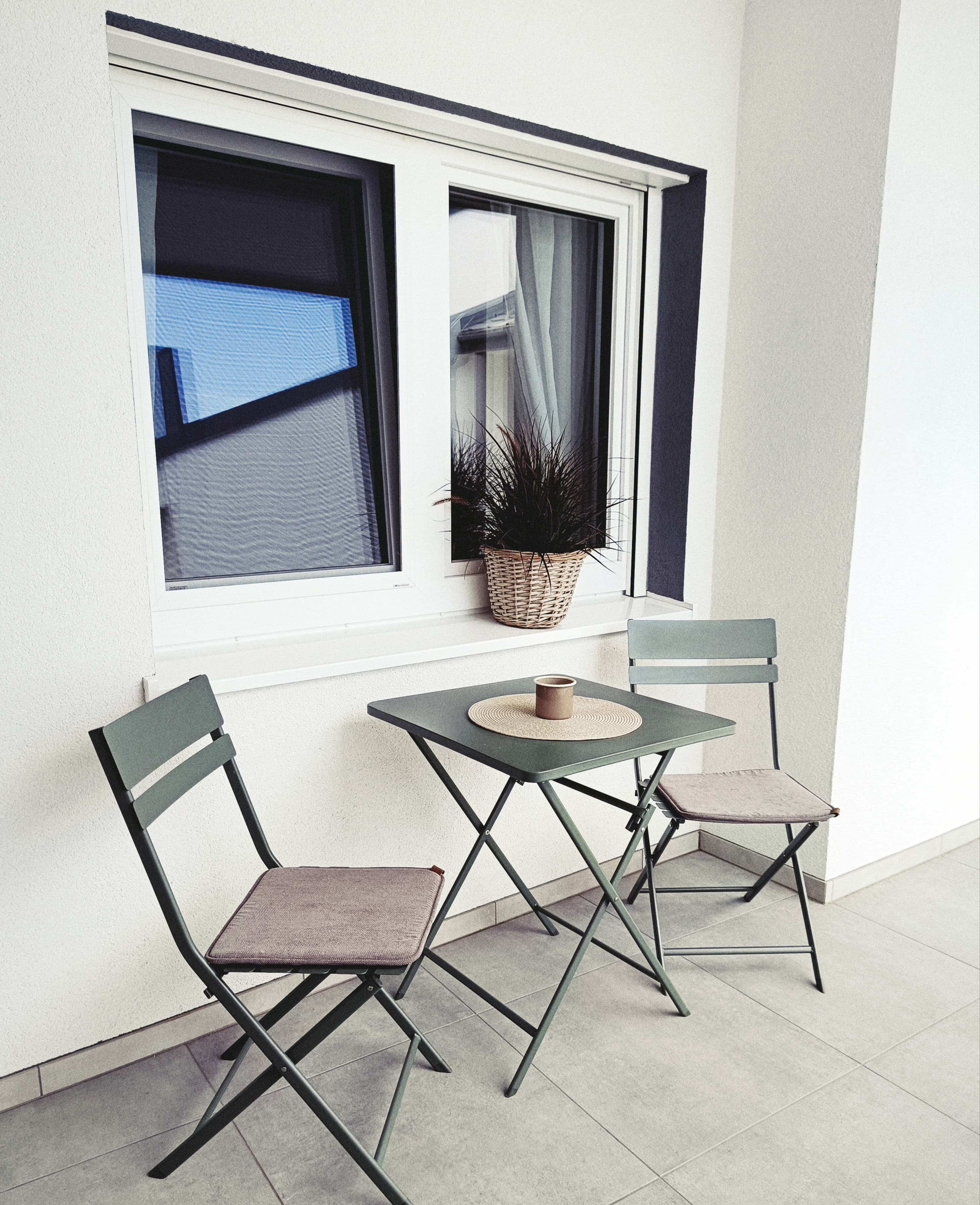 Kleines Balkon-Setup mit einem grünen Klapptisch und zwei passenden Stühlen auf hellen Fliesen. A potted plant sits on the window ledge, creating a serene vibe.