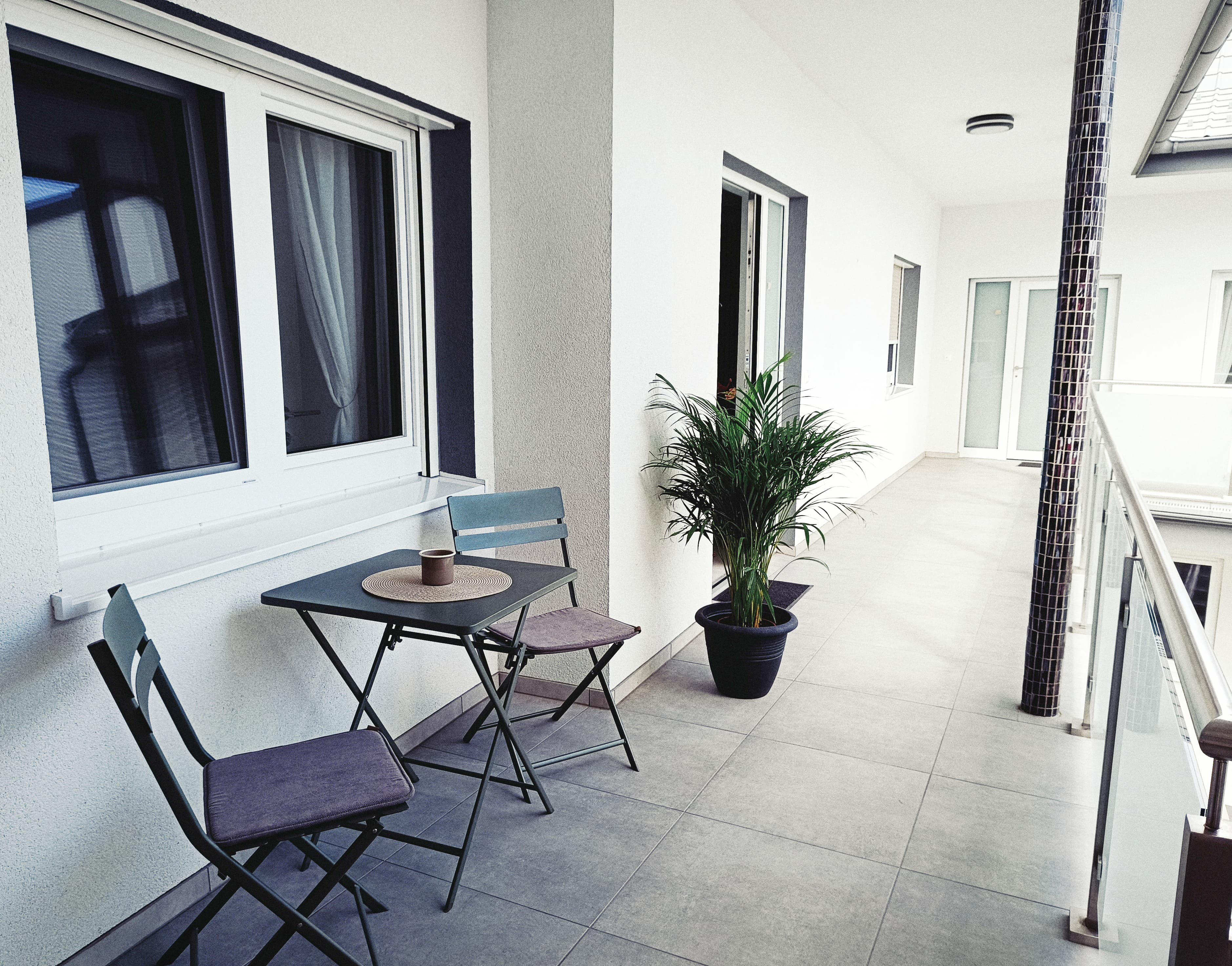 Moderner Balkon mit zwei grauen Klappstühlen, einem kleinen Tisch mit einer Tasse und einer Topfpflanze. Glass doors and railings create an airy feel.