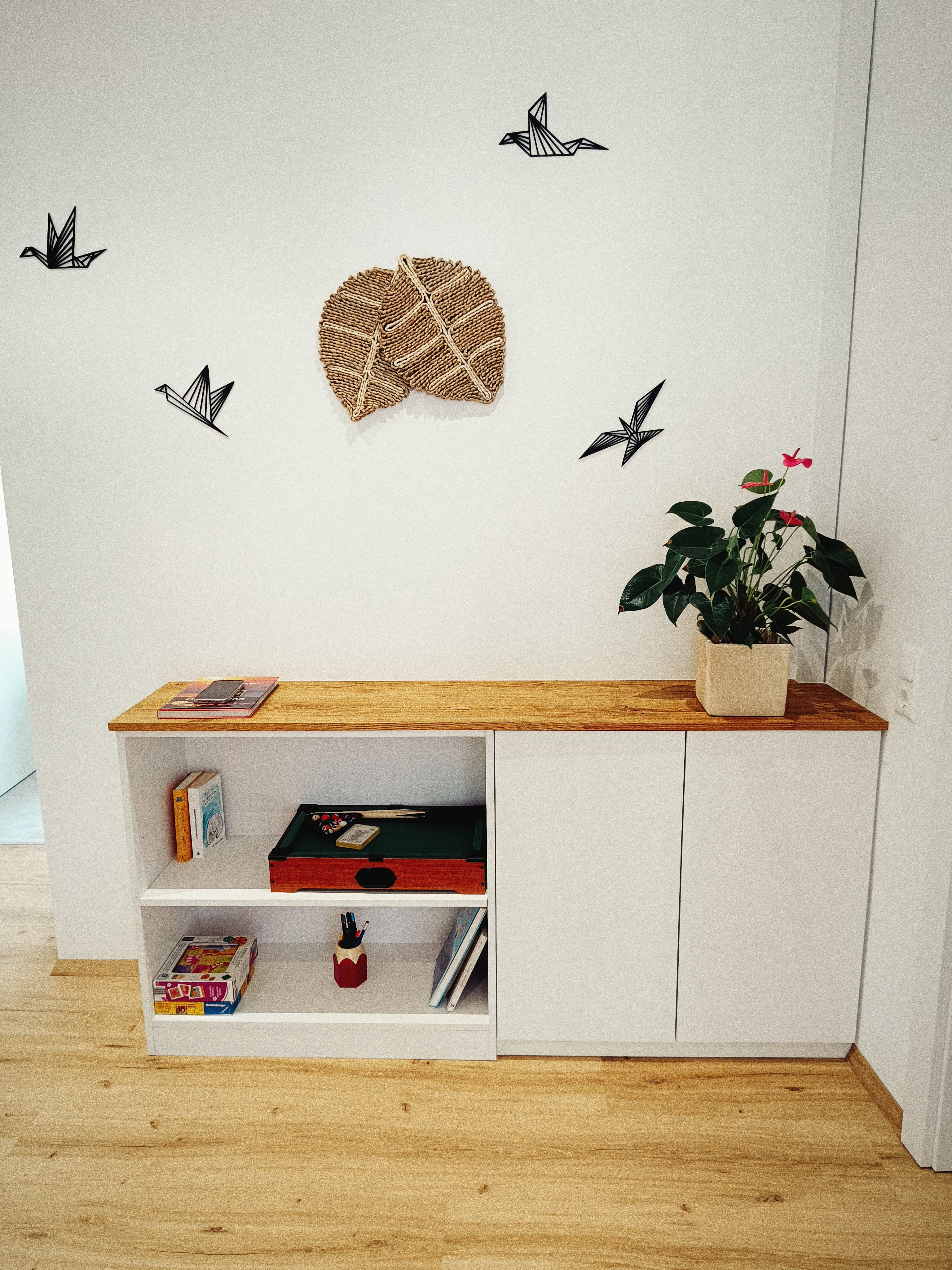 Moderner Wohnraum mit einem weißen Schrank, bekrönt mit einer Topfpflanze. Wall features artistic accents, including bird art and a textured centerpiece. Cozy and minimalist.