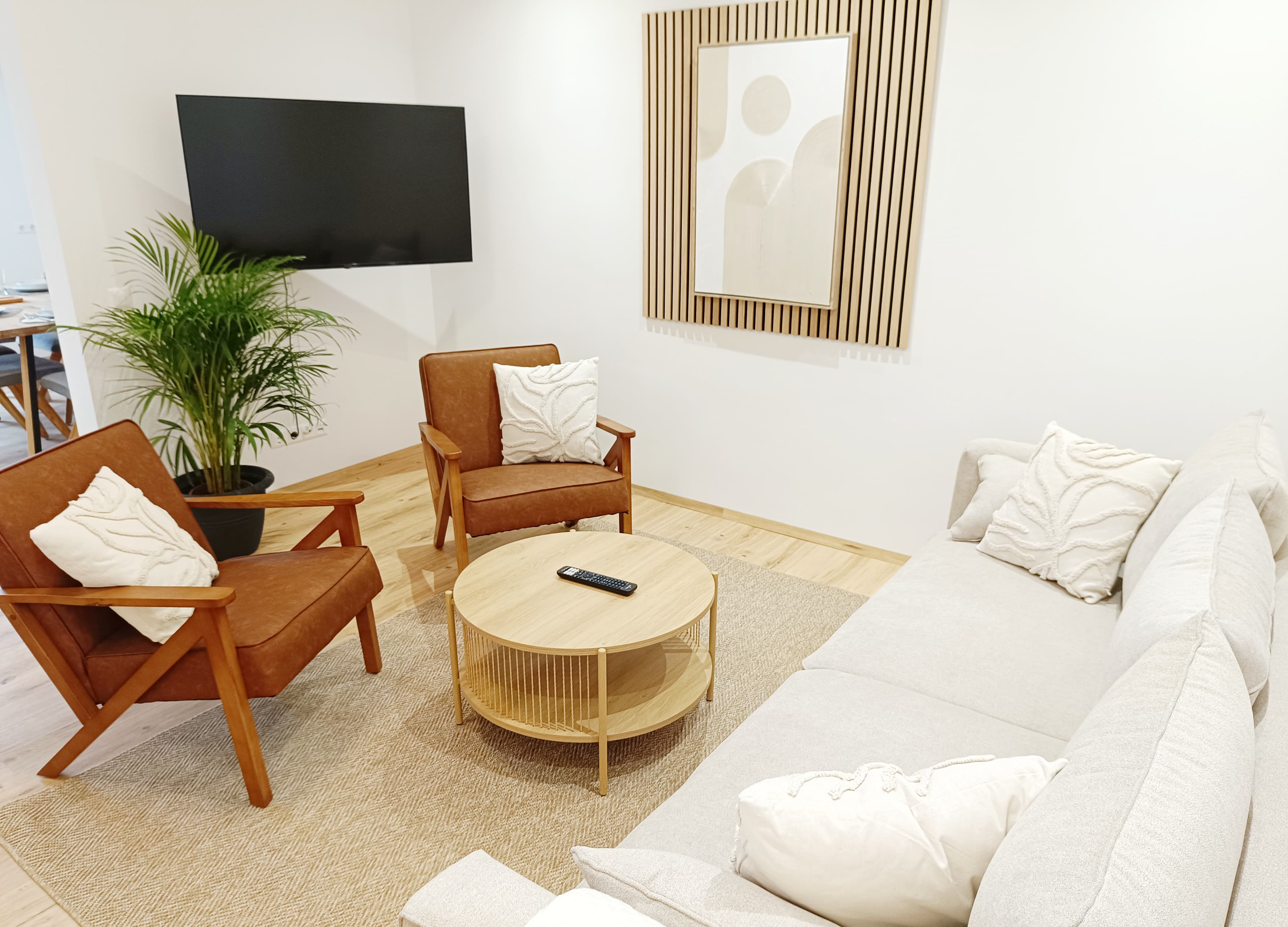 Modernes Wohnzimmer in neutralen Farbtönen, ausgestattet mit einem beigen Sofa, zwei braunen Sesseln, einem runden Couchtisch aus Weidengeflecht, Wandkunst, einer Zimmerpflanze und einem Fernseher.