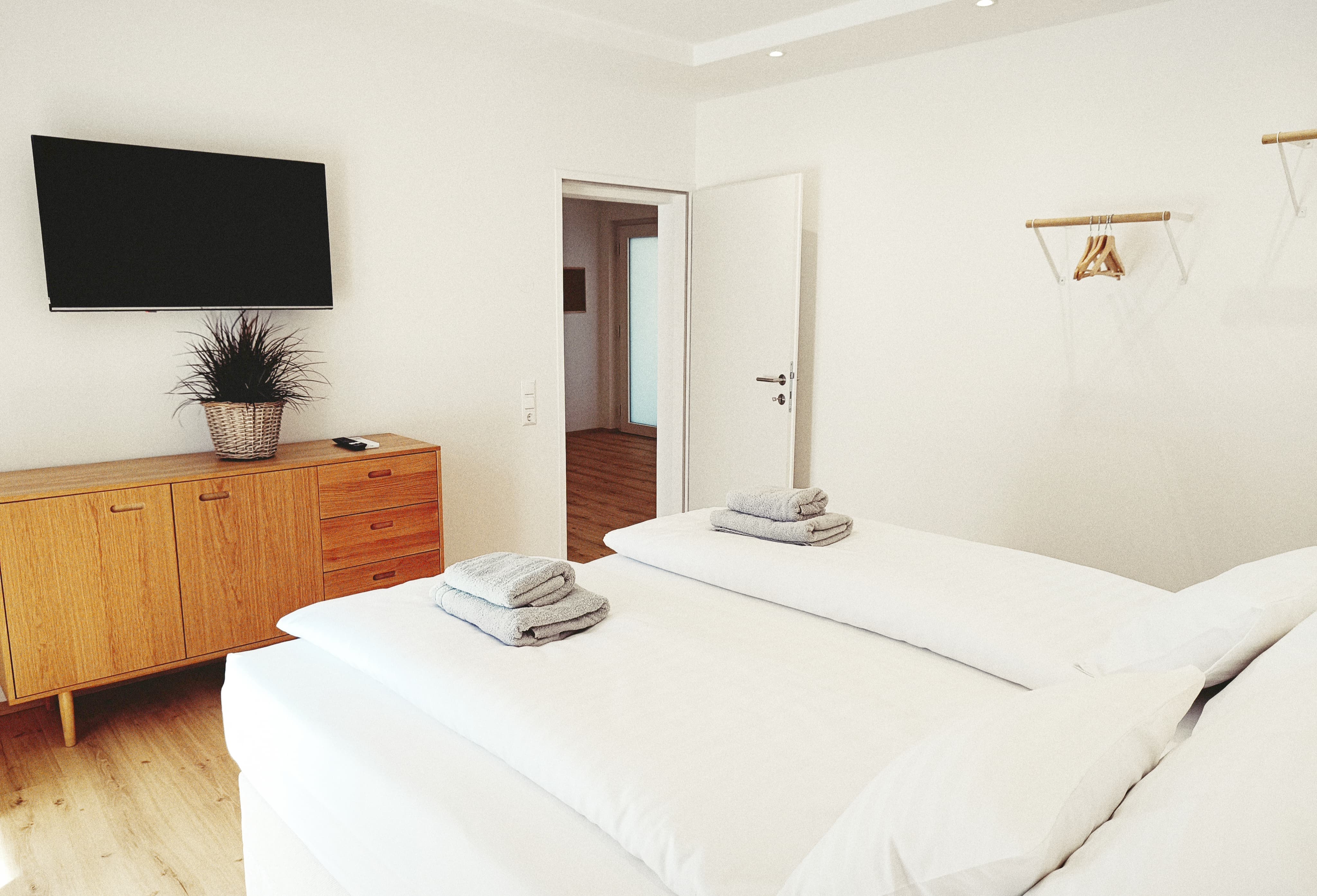 Modernes Schlafzimmer mit einem ordentlich gemachten Bett, gefalteten Handtüchern, einer Holzkommode, einem Wandfernseher und einer offenen Tür, die Hartholzboden zeigt. Calm and inviting atmosphere.