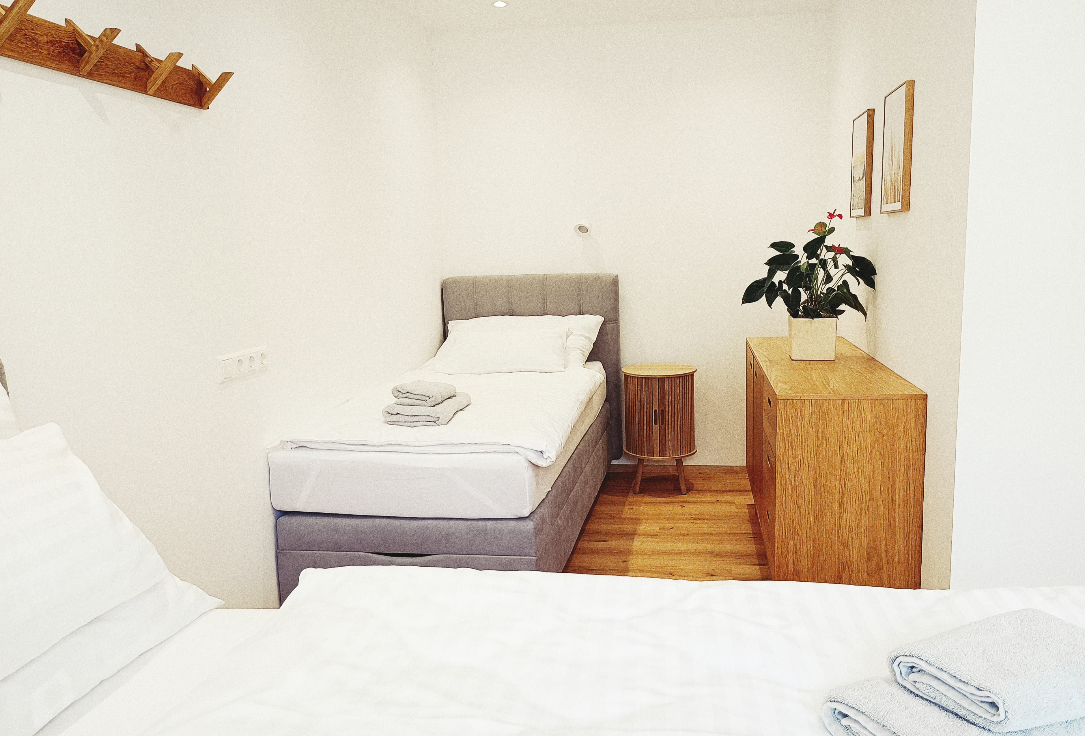 Gemütliches minimalistisches Schlafzimmer mit Einzelbett, grauem Kopfteil, Nachttisch aus Holz und Kommode. Plant, framed art, and soft lighting create a tranquil vibe.