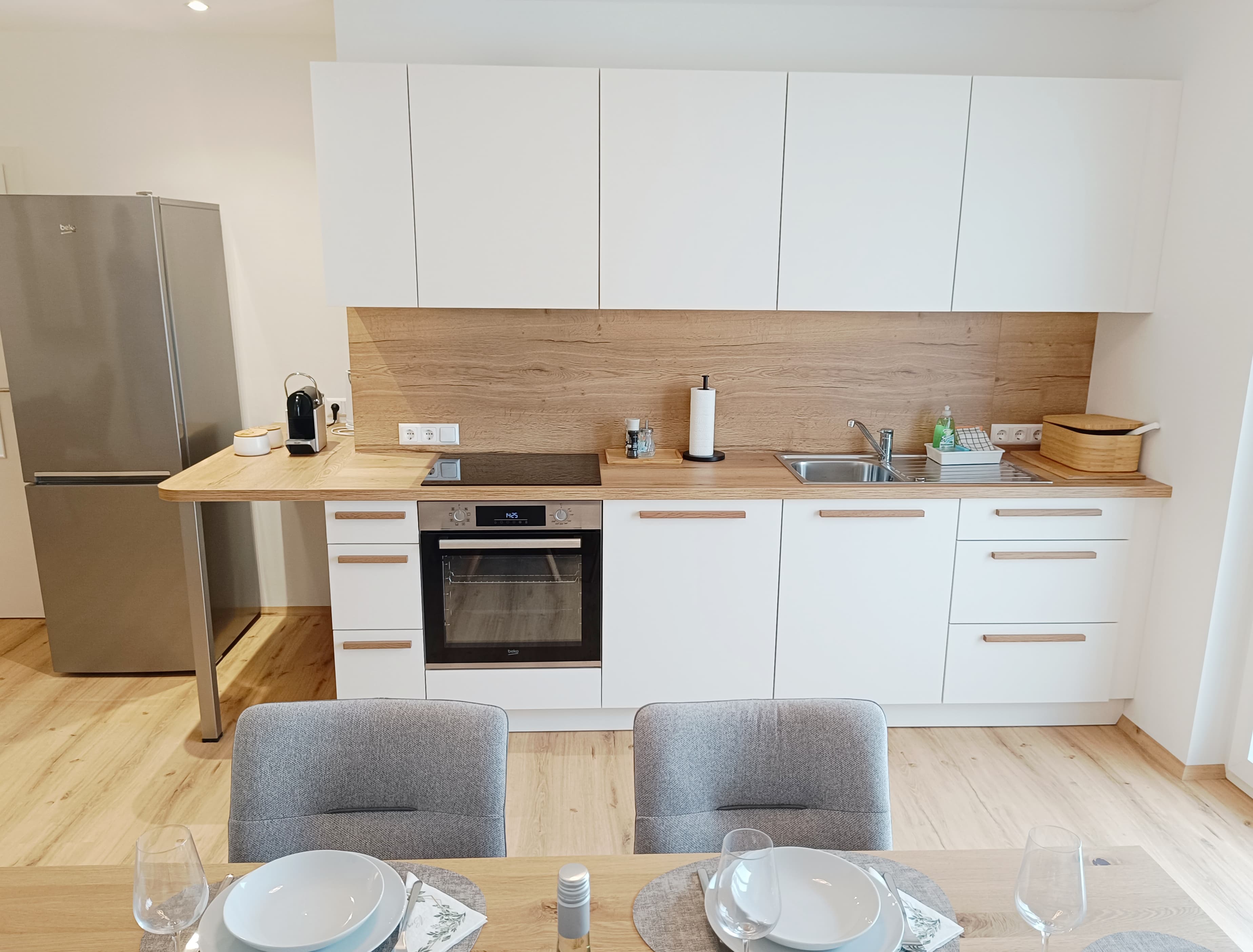 Moderne Küche mit weißen Schränken, Arbeitsplatten aus Holz und Rückwand. Stainless steel appliances, including a fridge and oven. Dining table set for four.
