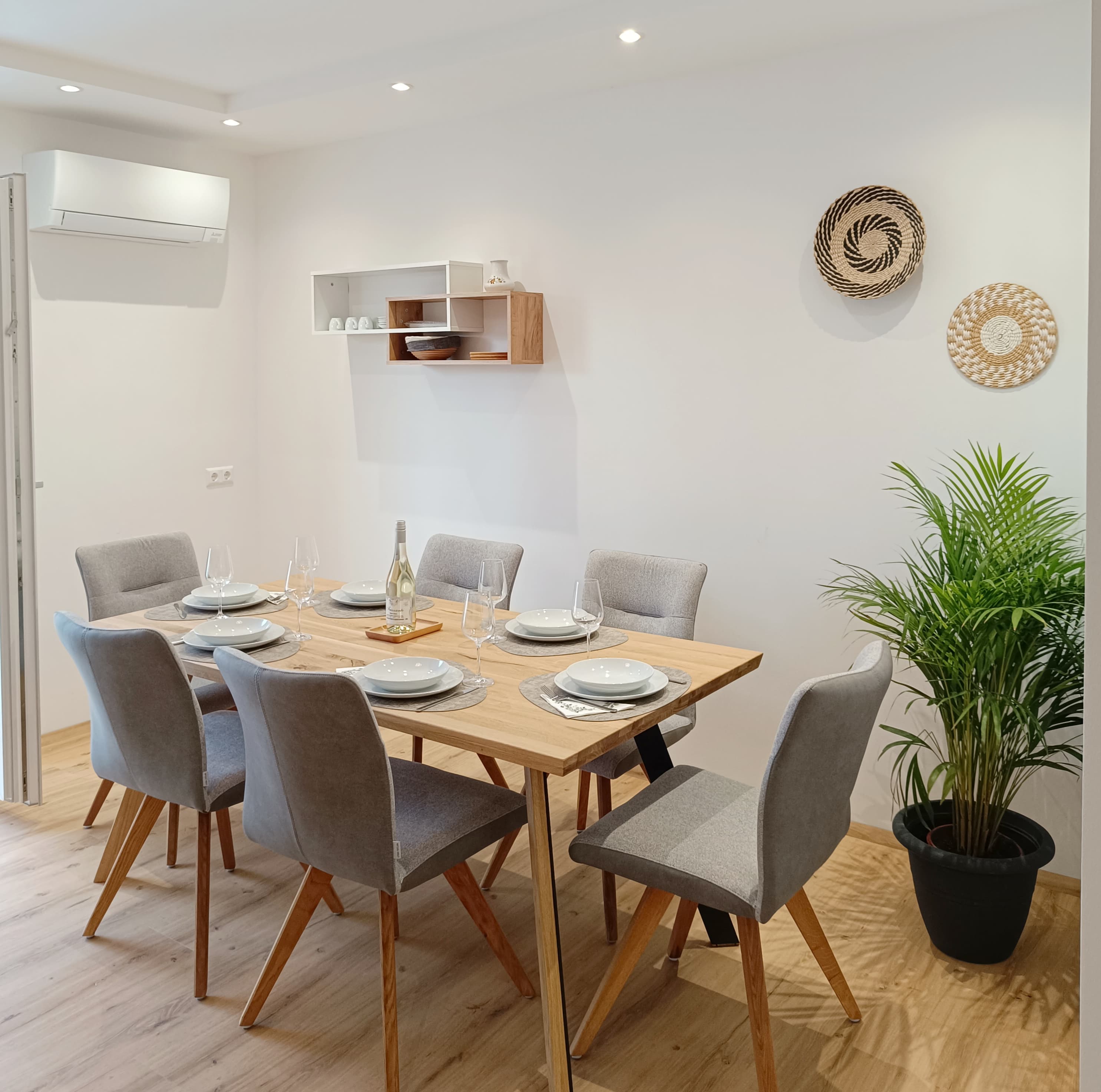 Modernes Esszimmer mit einem Holztisch für sechs Personen, ausgestattet mit grauen Stühlen und weißen Tellern. A potted plant and woven decor add a cozy touch.