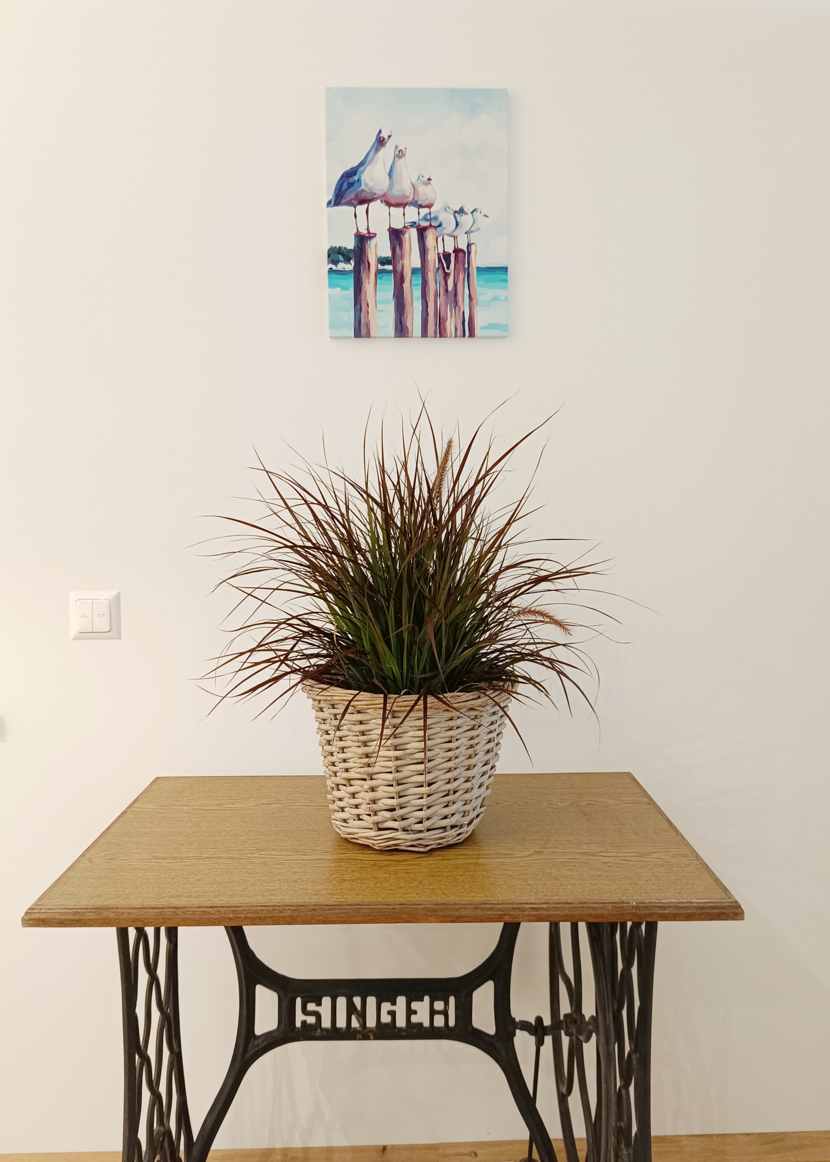 Eine Topfpflanze in einem geflochtenen Korb steht auf einem Vintage Singer Nähmaschinentisch vor einer weißen Wand. Above, a painting of seagulls on posts with a sea backdrop.
