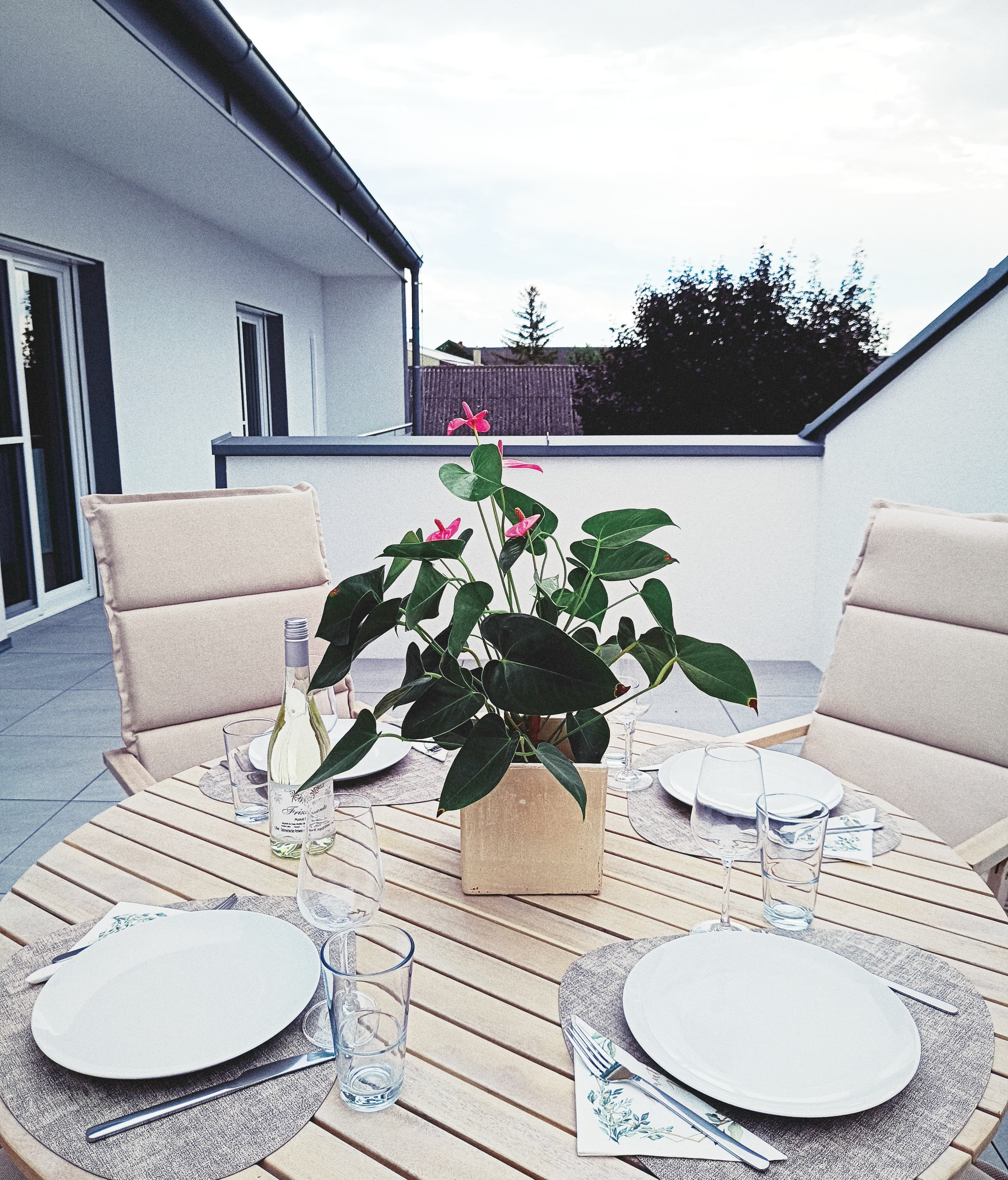 Außenbereich mit einem runden Holztisch für vier Personen, in der Mitte ein Topf mit einer Pflanze. Soft beige chairs, wine bottle, and glasses add a cozy ambiance.