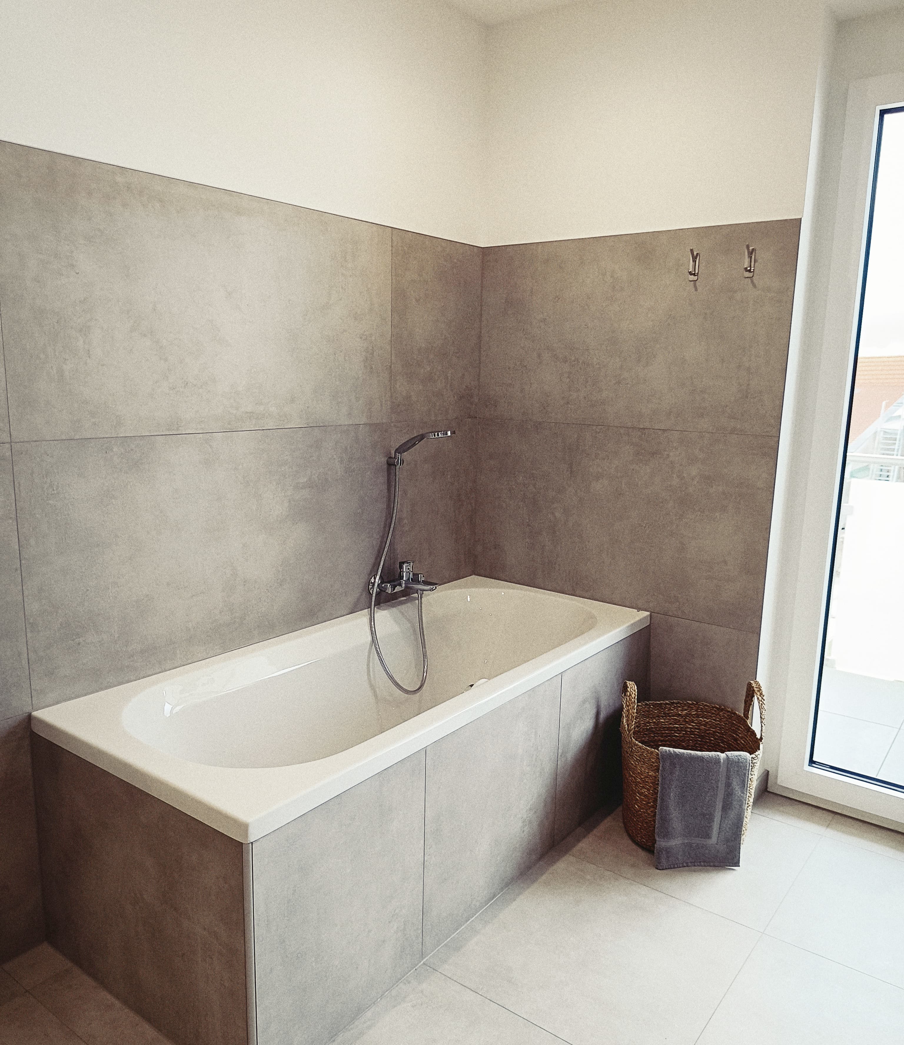 Modernes Badezimmer mit einer eleganten Badewanne, grauen Fliesen, einer Duschkopf-Armatur und einem geflochtenen Korb mit einem gefalteten Handtuch, was eine saubere, minimalistische Atmosphäre vermittelt.