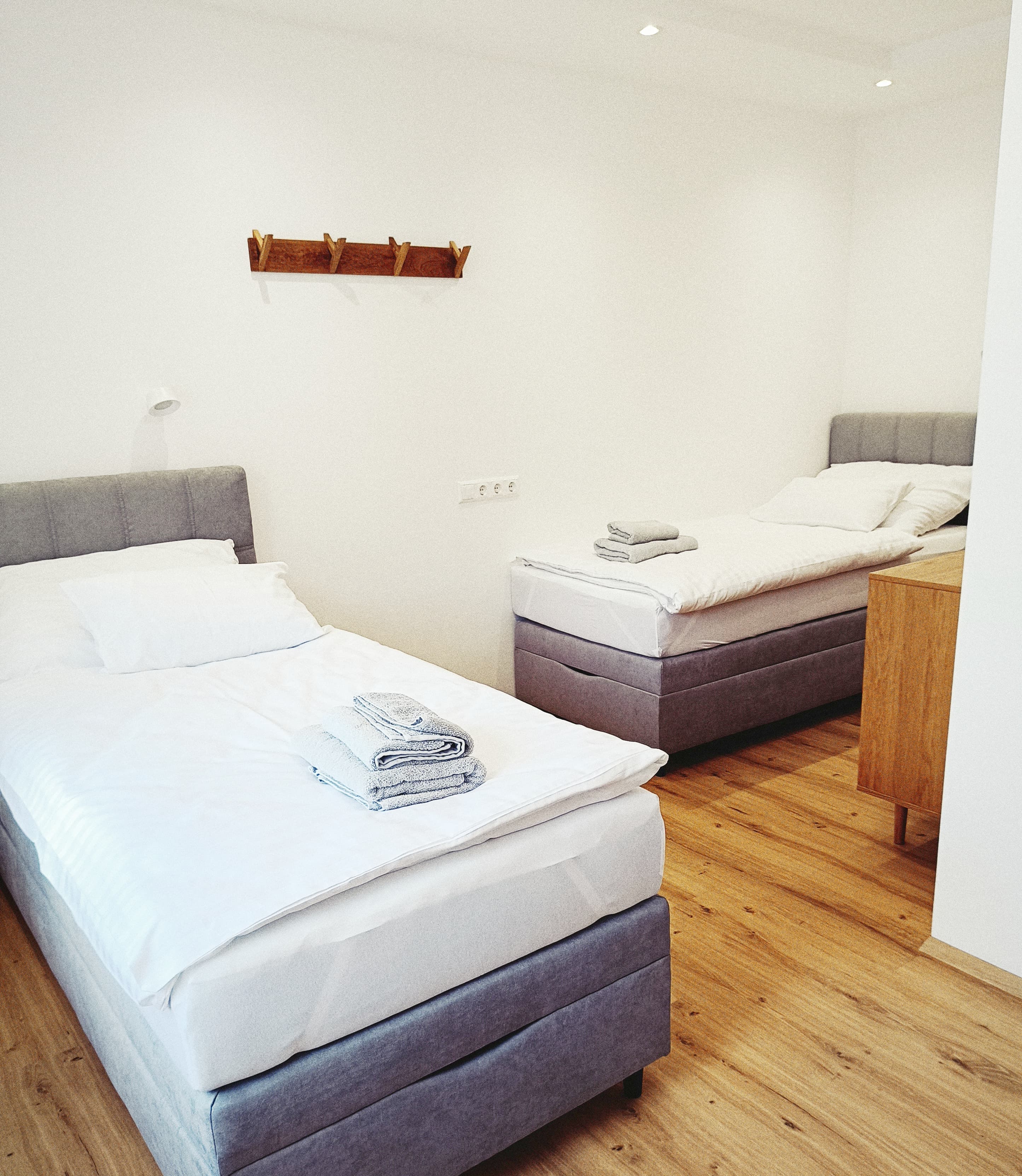 Zwei ordentlich gemachte Einzelbetten mit weißer Bettwäsche und gefalteten Handtüchern in einem hellen, minimalistischen Raum. Wooden floor and coat rack enhance the cozy ambiance.